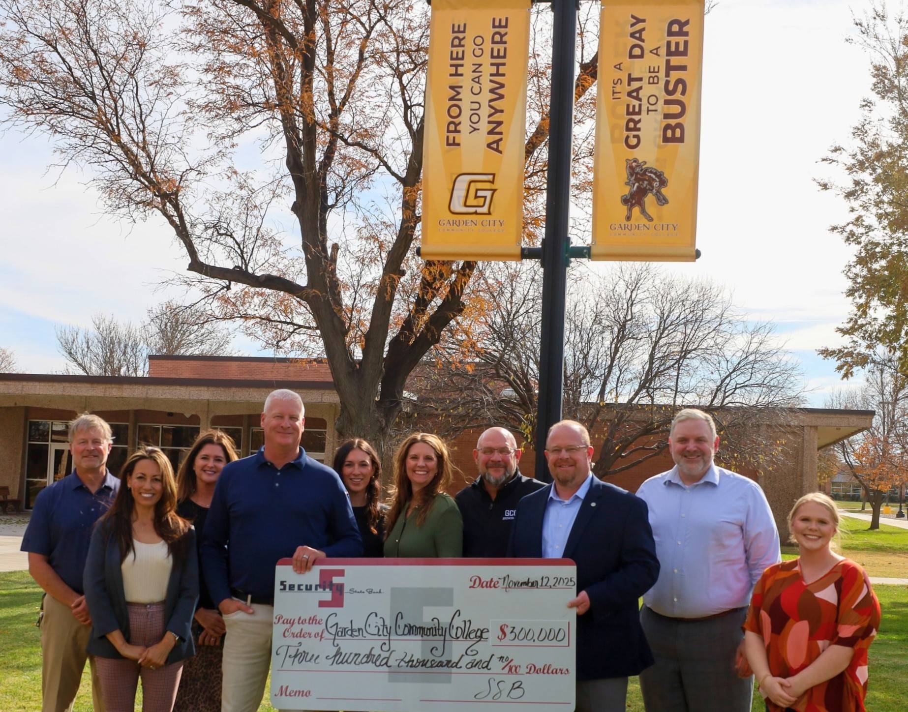 Group holding a large ceremonial check to Garden City Community College, standing outdoors near campus banners and trees.