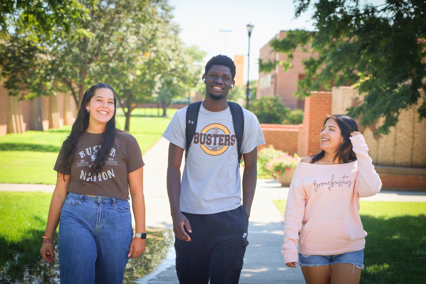 Three people walking together on a green campus walkway, wearing casual clothes and backpacks.