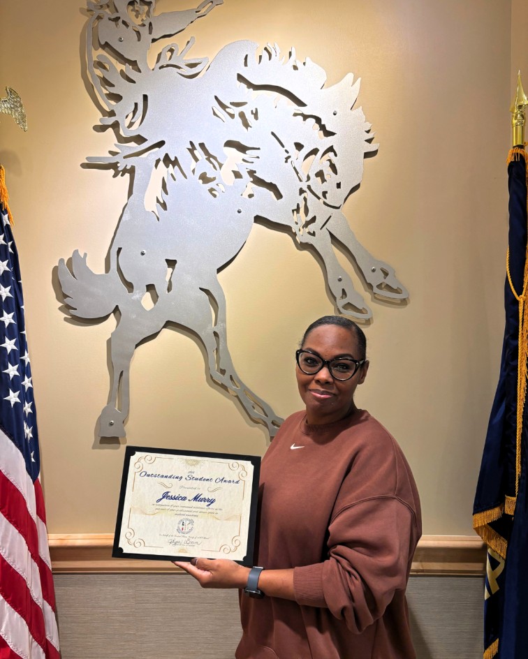 Woman holding an Outstanding Student Award certificate, standing between American and state flags with a large metal horse and rider sculpture behind her.