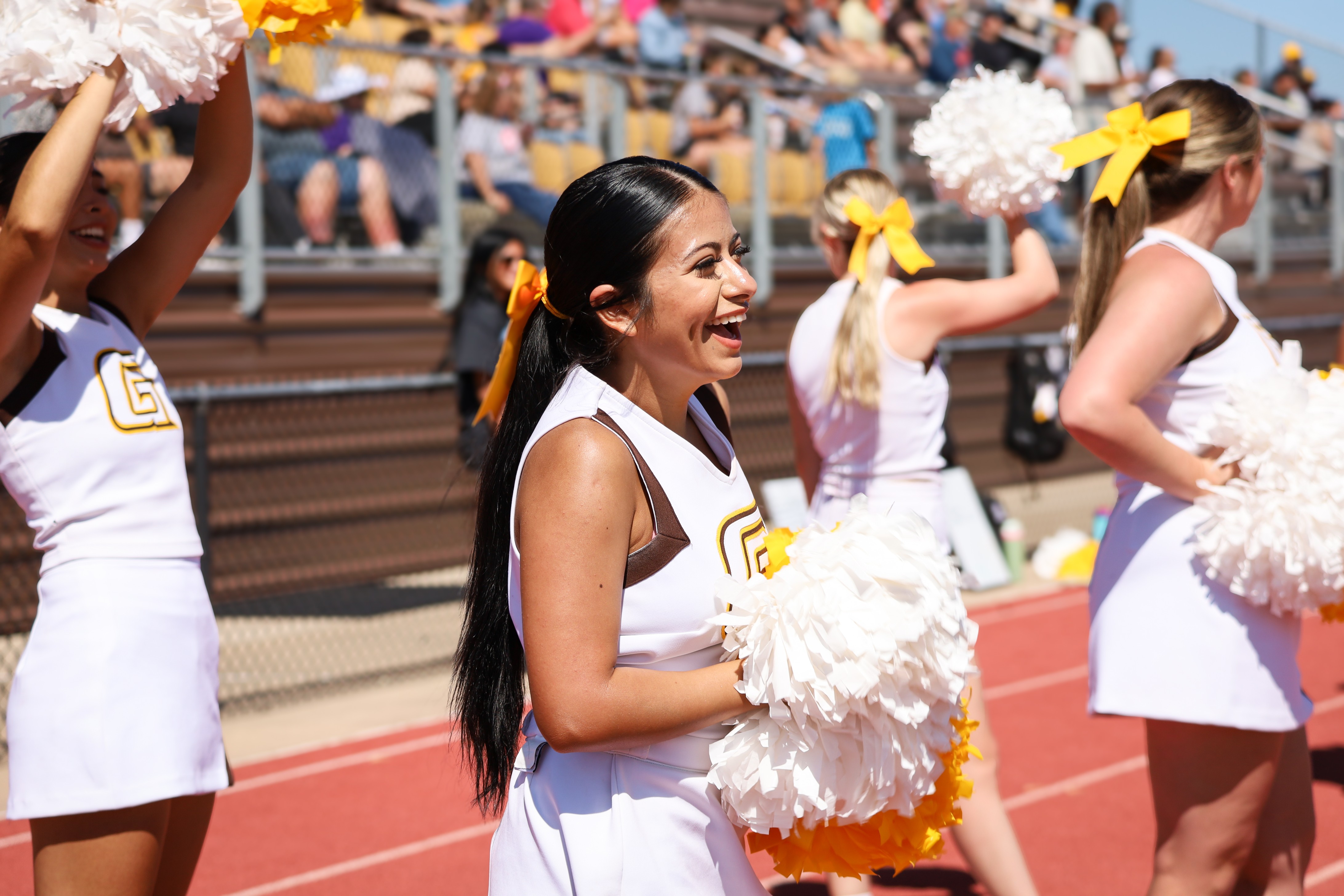Cheerleaders performing on the sidelines of a college athletic event, holding pom-poms in front of stadium seating.