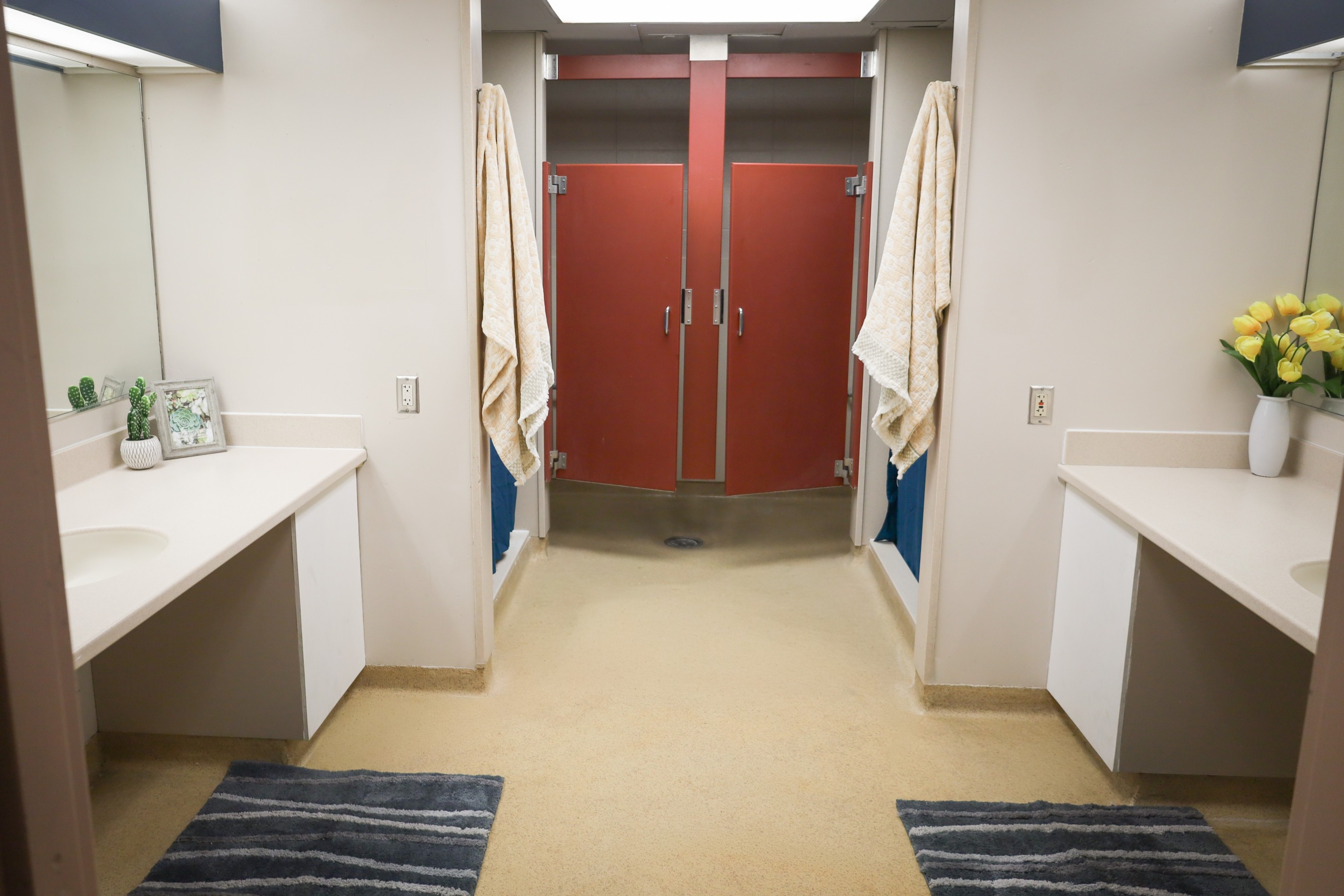 Shared bathroom area with two sinks and mirrors, bath mats on the floor, and shower stalls with red partition doors and hanging towels.