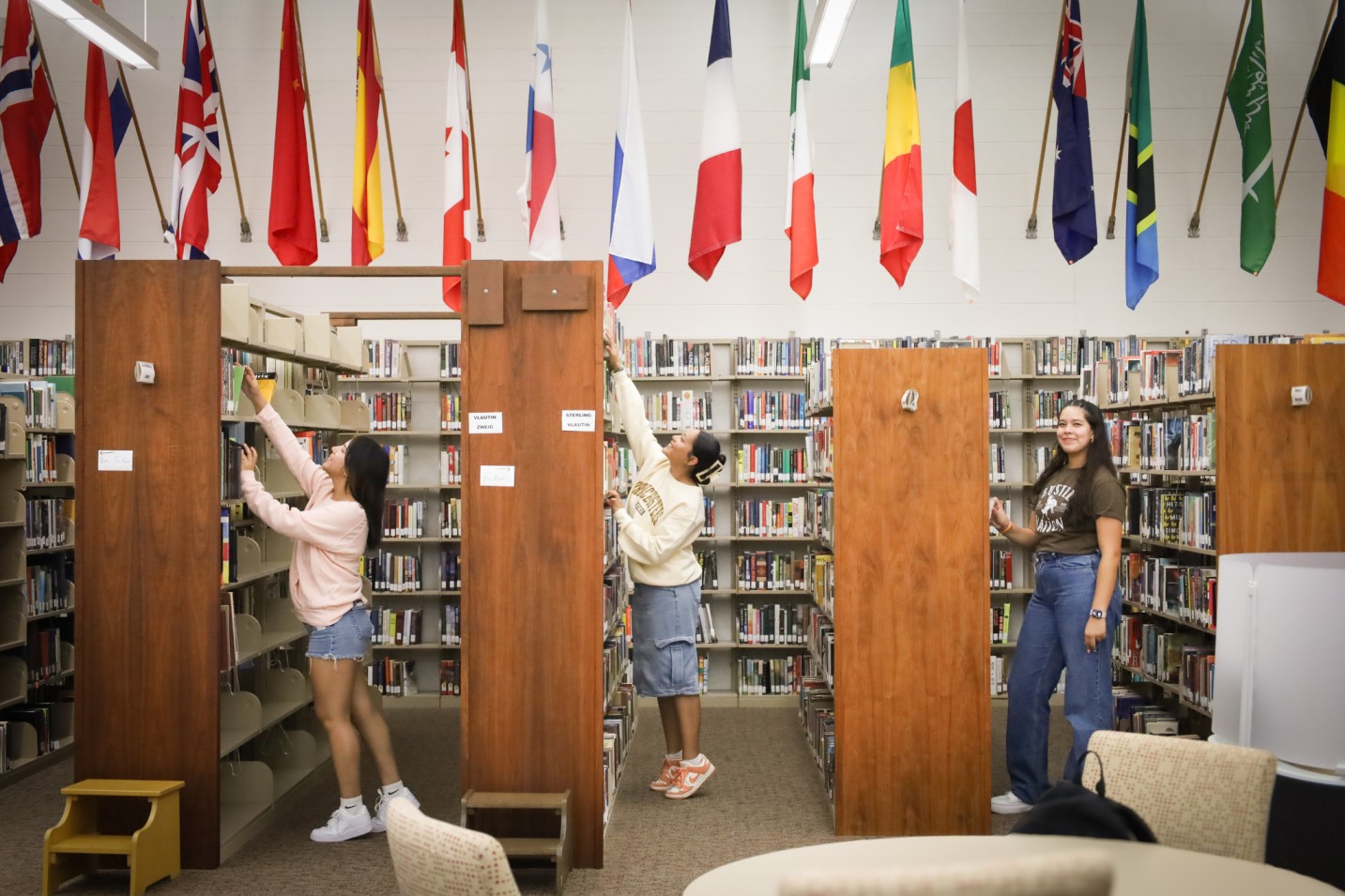 Copilot said:  Students browsing and selecting books from library shelves beneath rows of international flags.
